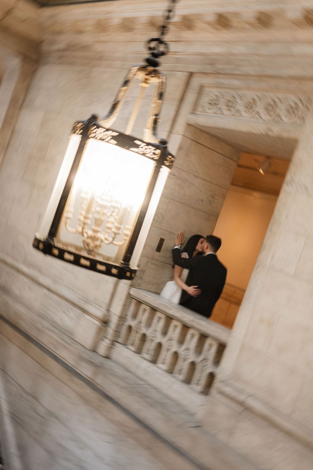 Couple sharing a kiss on a grand marble balcony, reflected in a ornate hanging lantern at an elegant wedding venue.