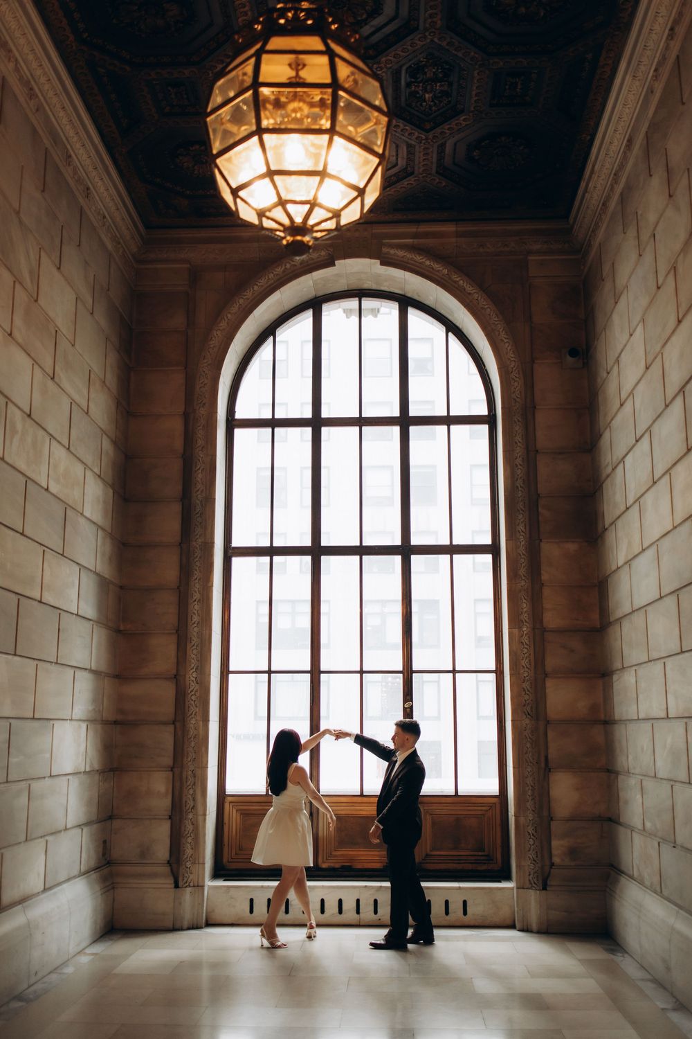Couple dancing elegantly in a grand hall with arched window, ornate ceiling, and vintage chandelier.
