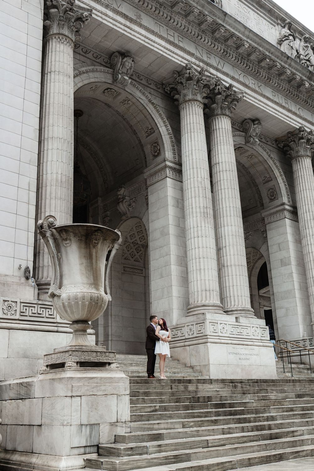 Couple embracing on steps of New York Public Library with grand columns and stone urn.