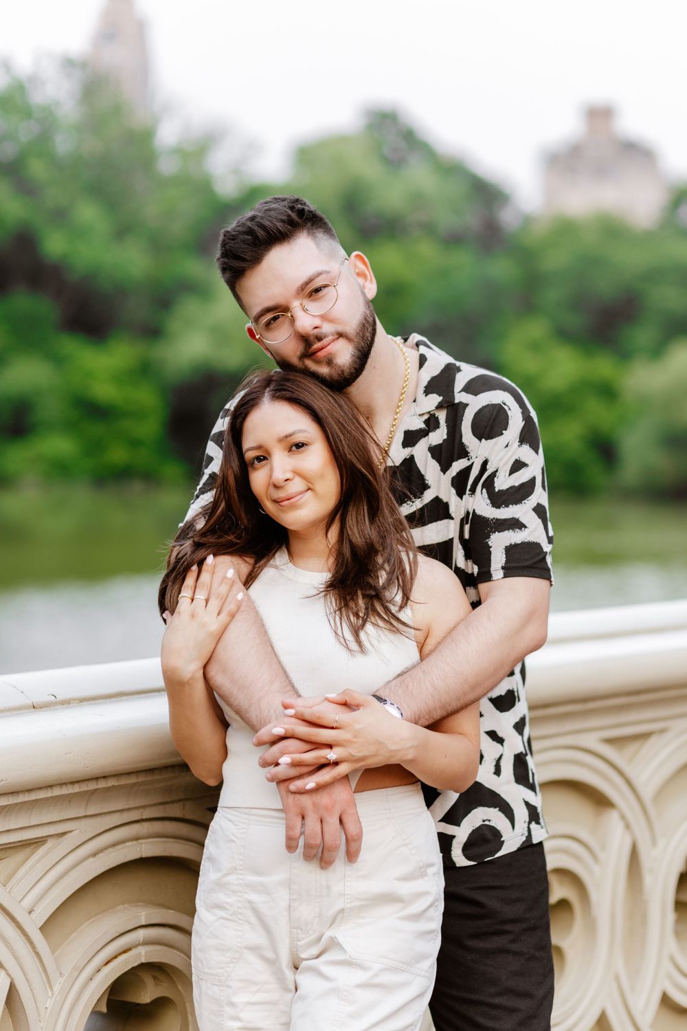 A couple embraces on a stone bridge overlooking a lake, with lush greenery in the background on a sunny day.