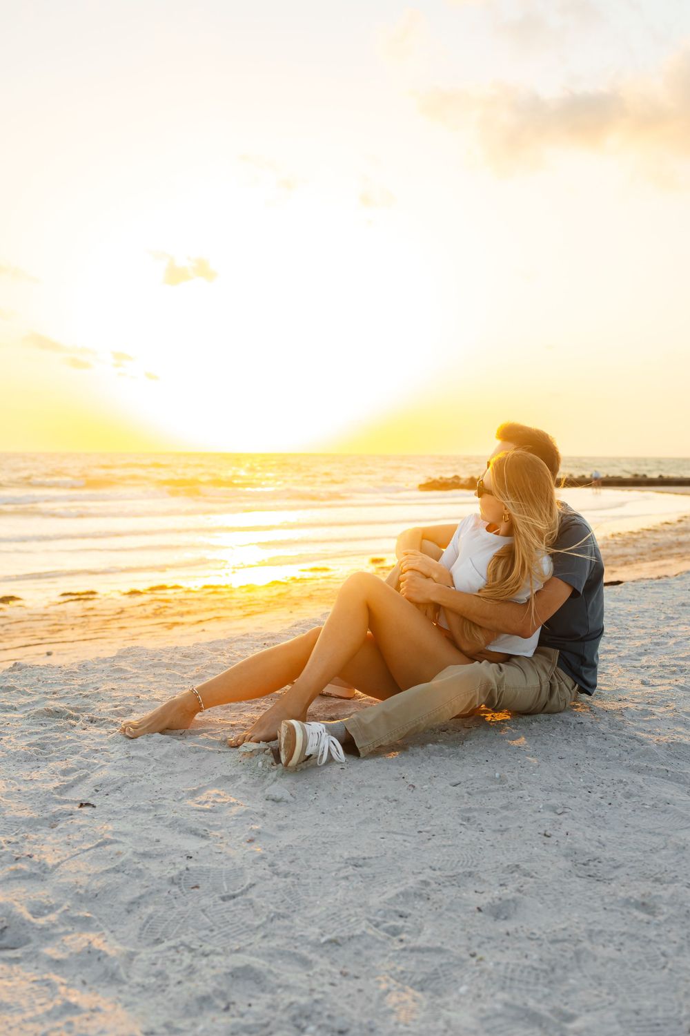 A couple relaxes on a sandy beach at sunset, cuddling together as golden sunlight illuminates the shoreline.