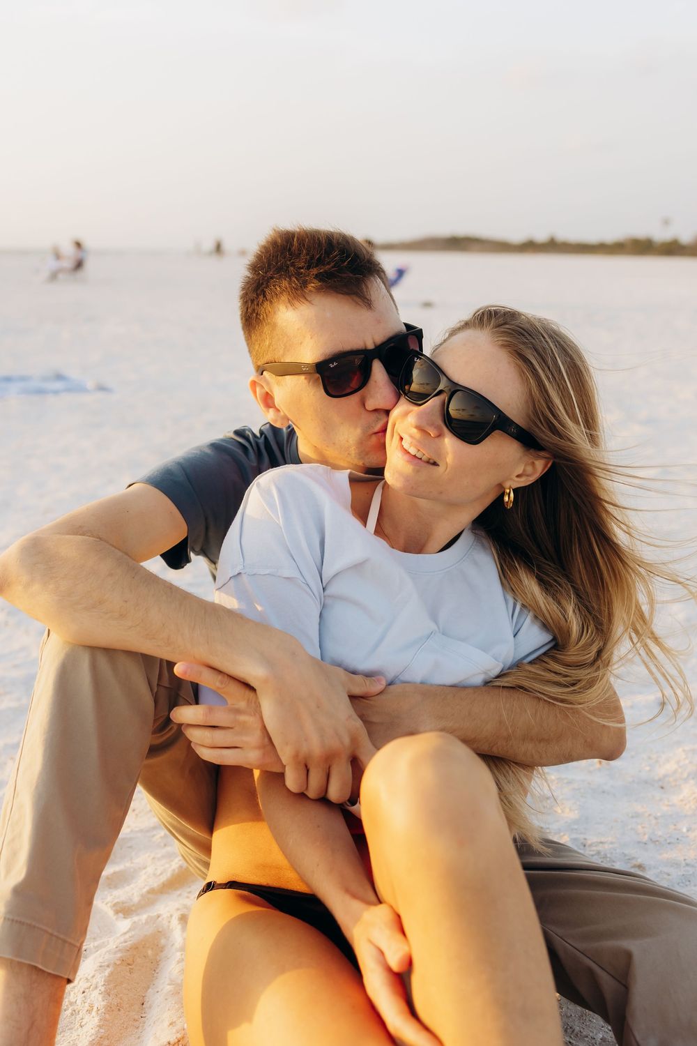 A couple wearing sunglasses embraces on a white sandy beach during golden hour.