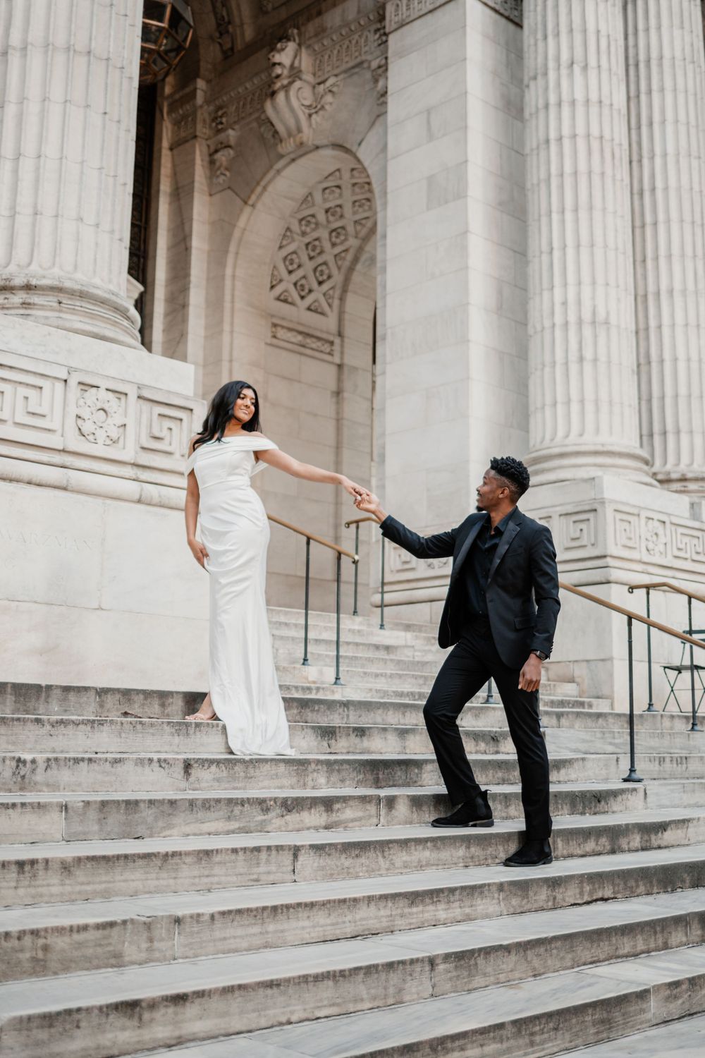 Elegant couple in formal attire pose on grand marble steps of a classical building with columns.