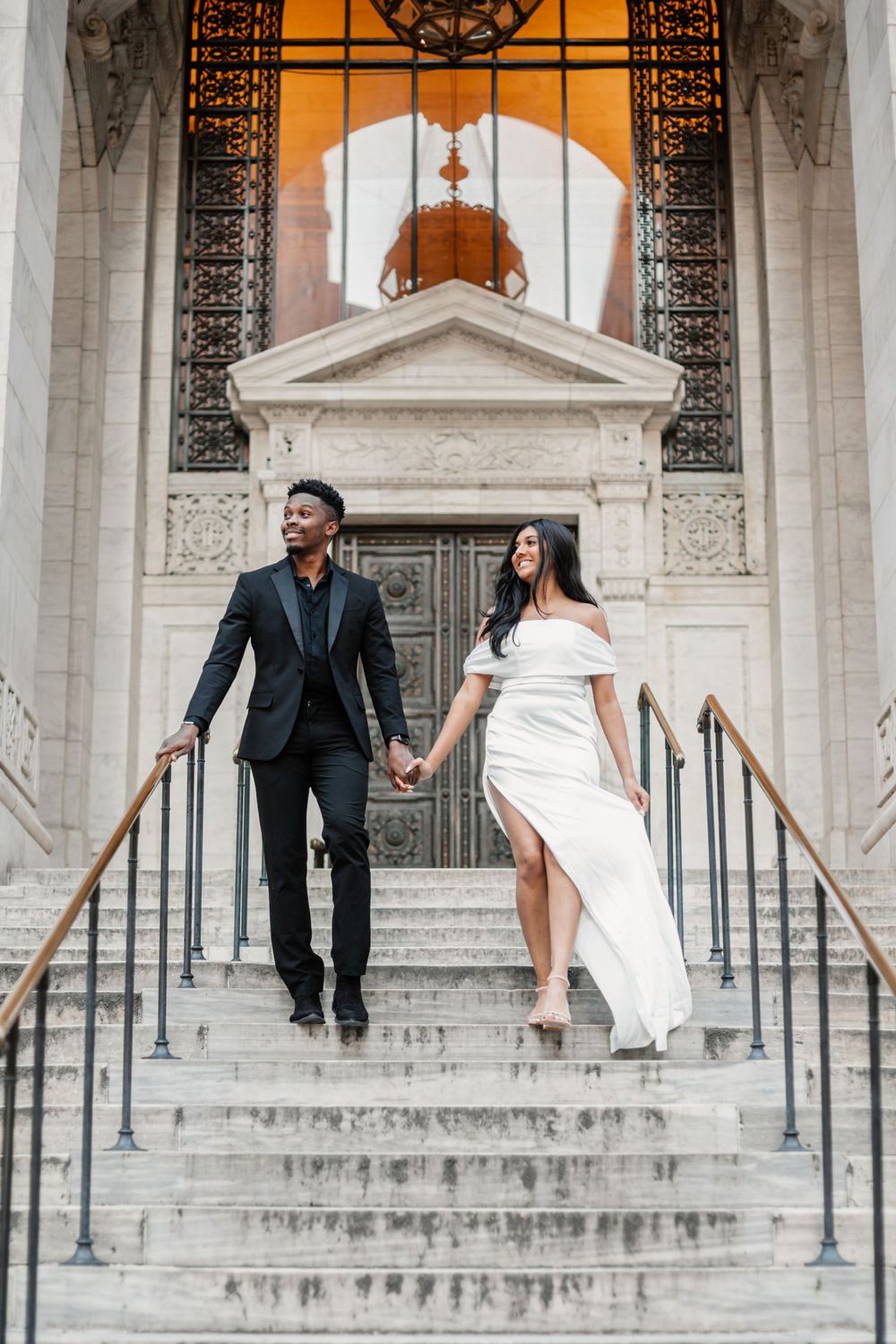 A couple descends grand stone steps of a historic building as the bride wears a flowing white dress and groom in dark suit.