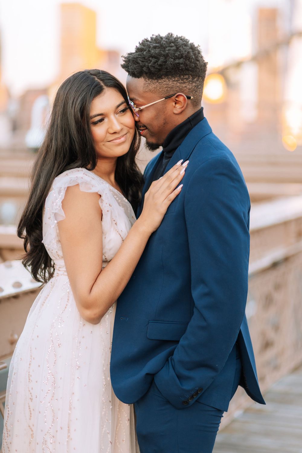 A romantic couple embraces outdoors, with her in a white lace dress and him in a navy blue suit.