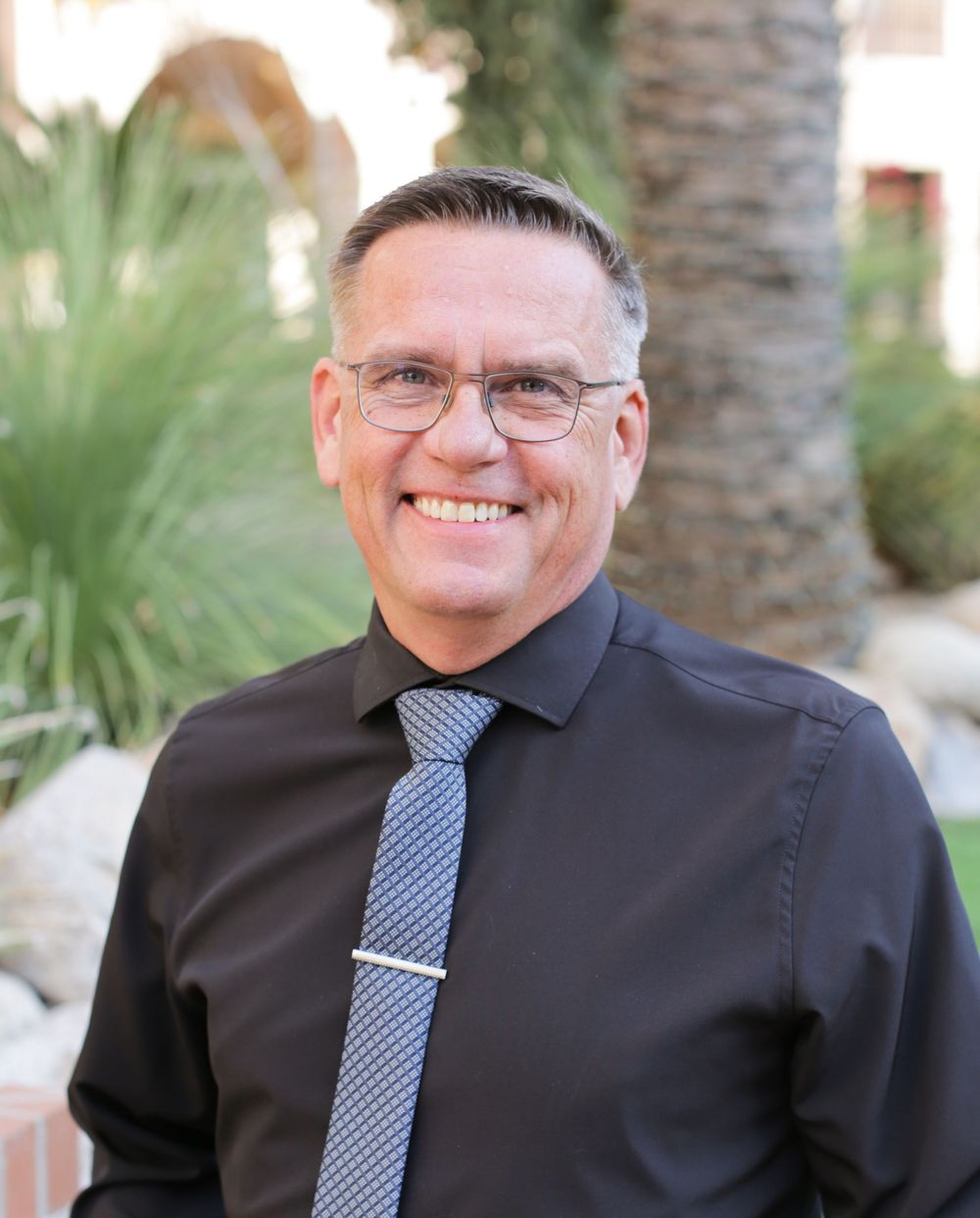 Professional headshot of smiling man in black with tie and desert background