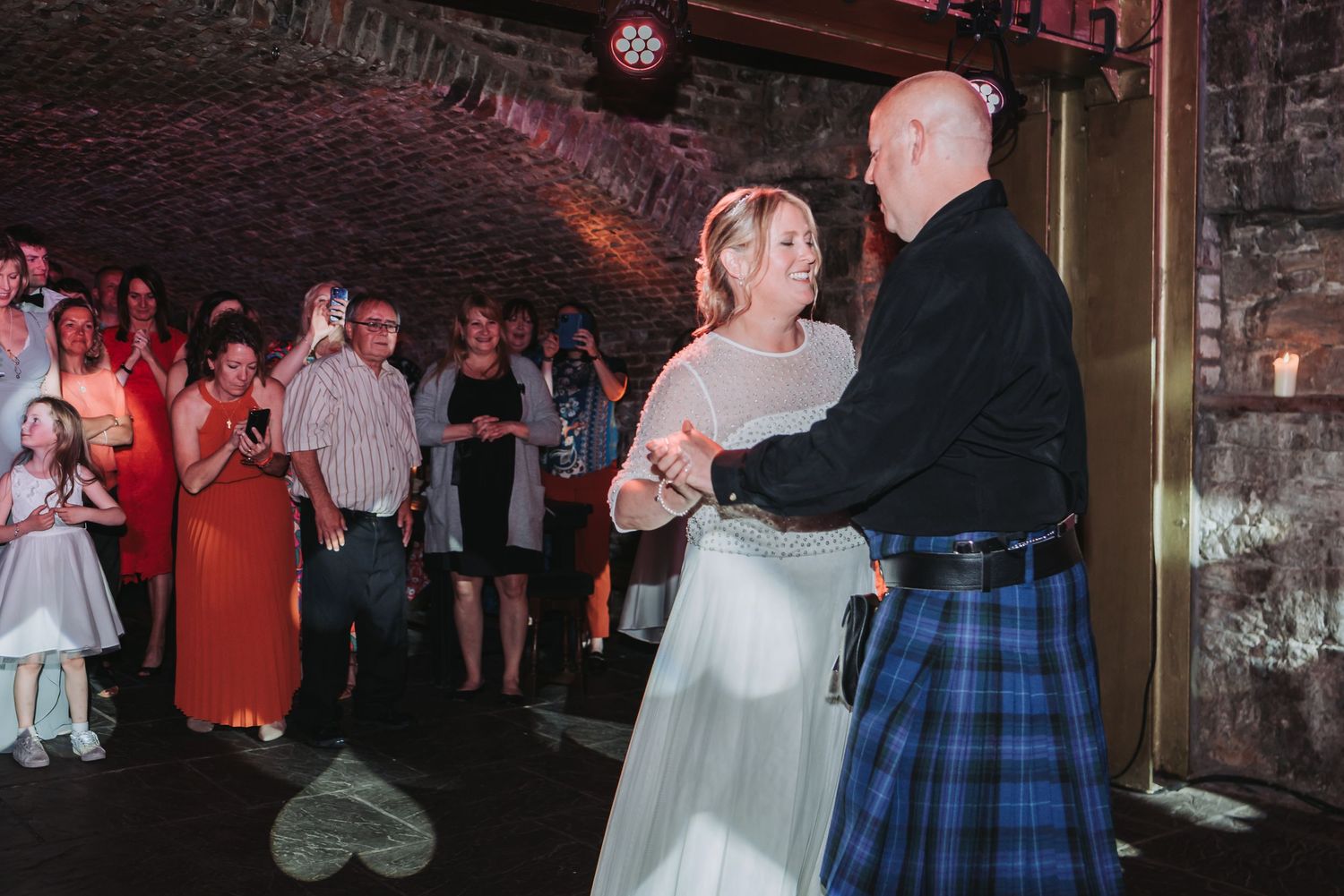 A couple sharing their first dance in a romantically lit underground venue with guests watching.