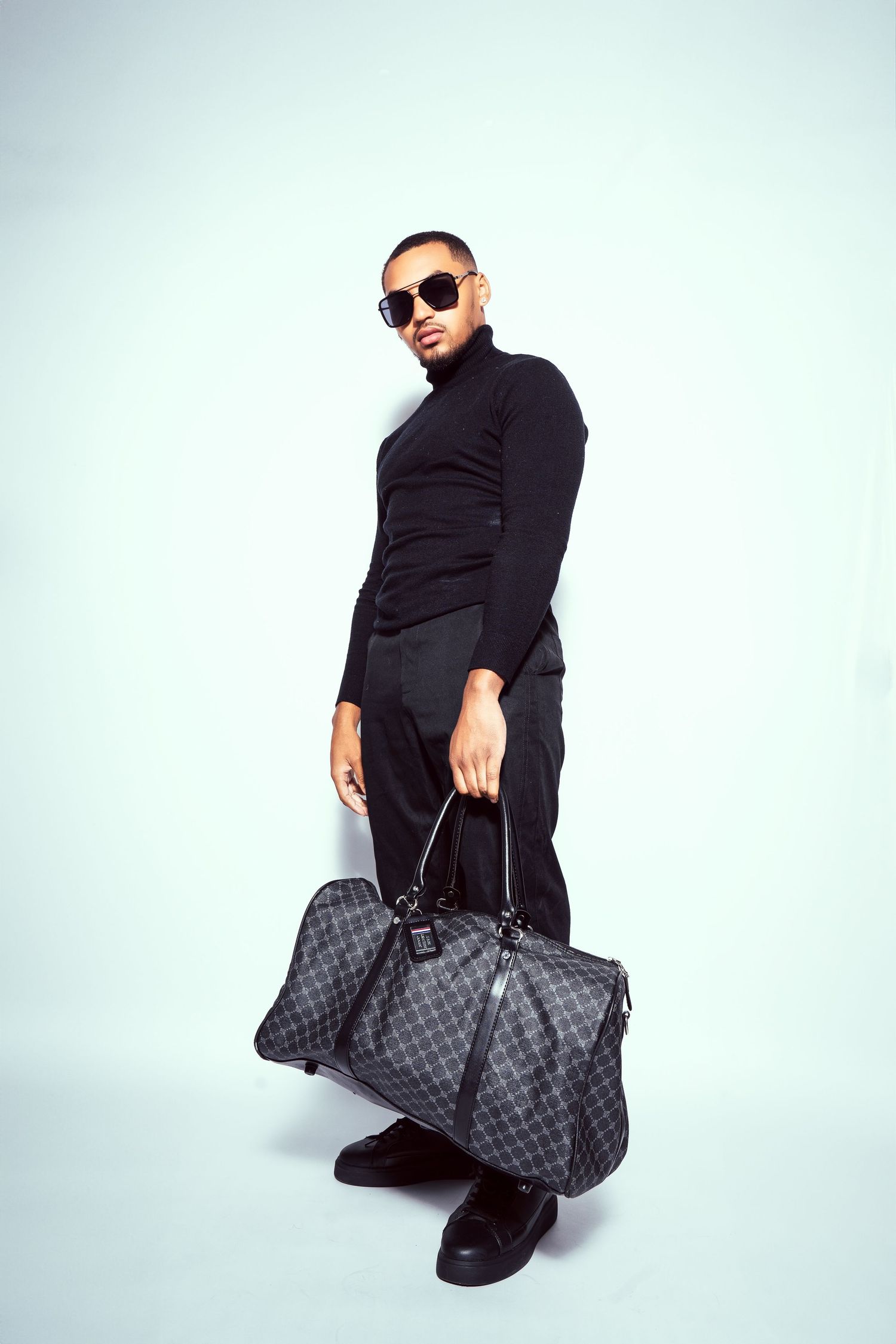Stylish model wearing black turtleneck and sunglasses holding designer travel bag.
