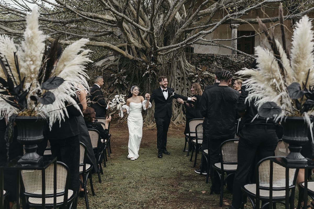 Bride and groom walk down outdoor wedding aisle lined with pampas grass arrangements and guests in black attire.