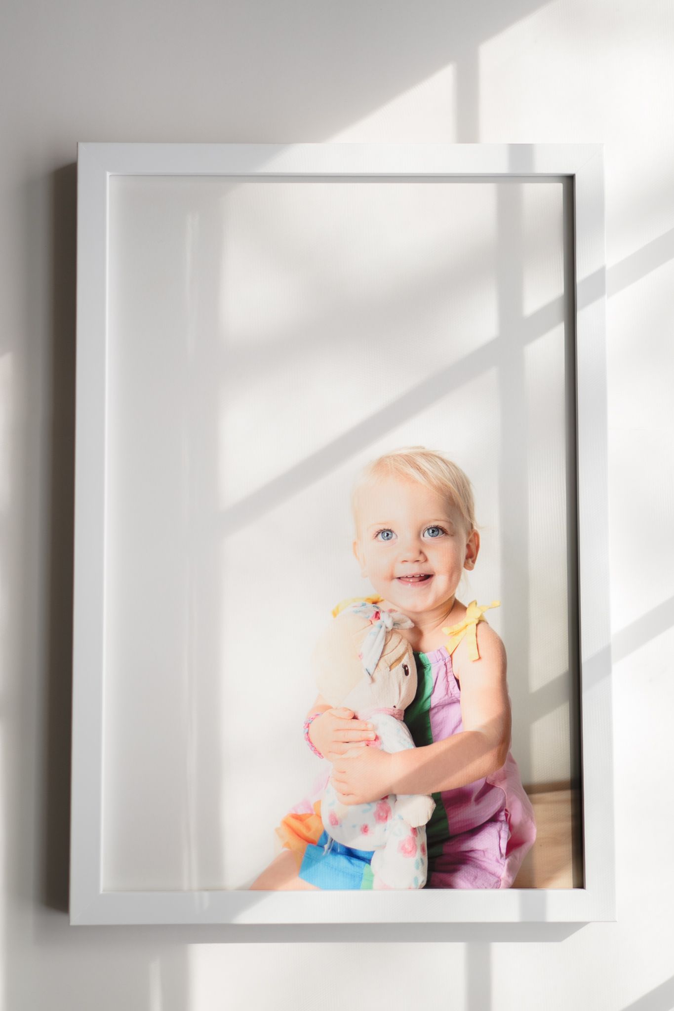 Framed portrait photograph showing a young child in pink clothing smiling against a white wall with natural window light.