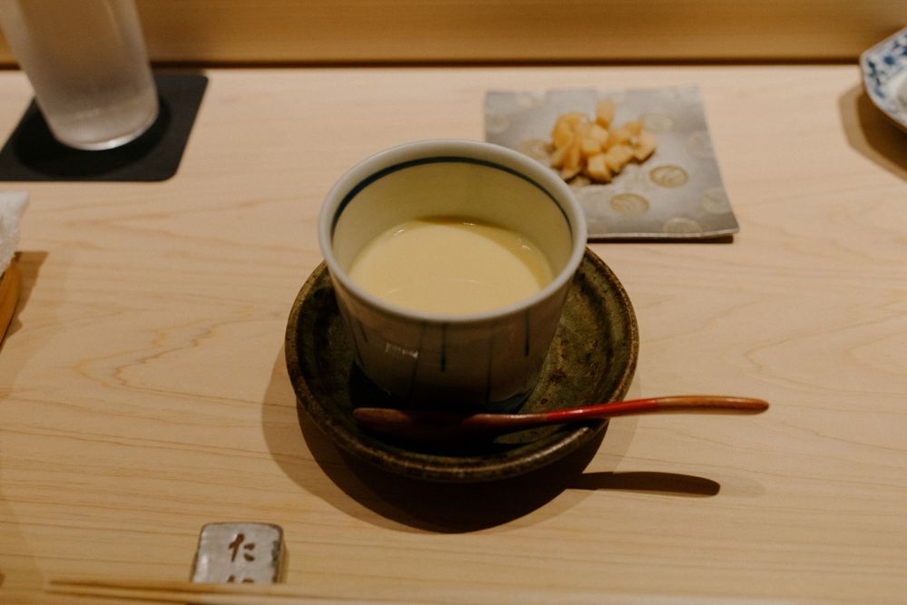 A traditional chawanmushi egg custard served in a dark ceramic bowl with wooden spoon.