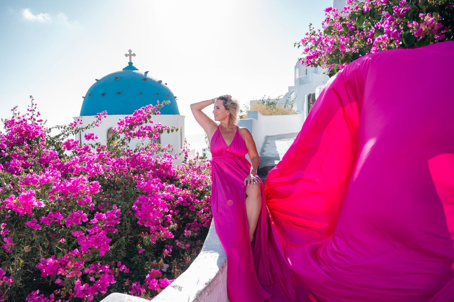 Woman in flowing pink dress poses among vibrant bougainvillea flowers with Greek church dome in background.