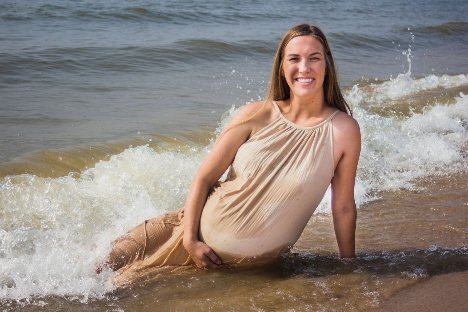 Maternity photo of a woman holding her baby bump with a wave crashing over her legs.