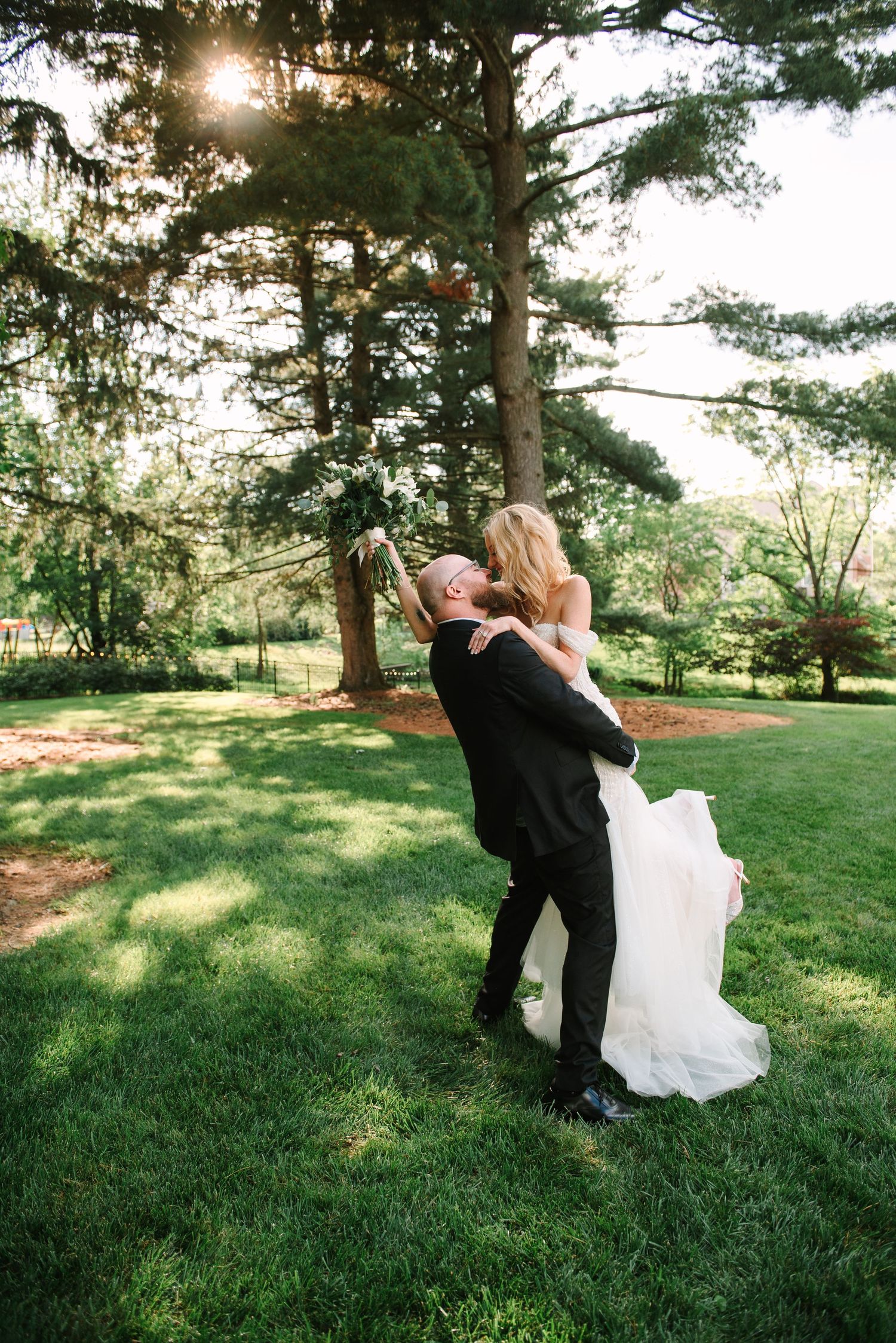 Joyful wedding couple embracing on sunny lawn with streaming sunlight through pine trees.