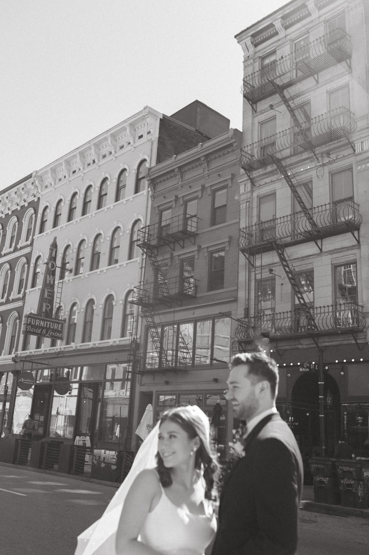 A wedding couple poses on a city street with classic architecture and fire escapes in the background.