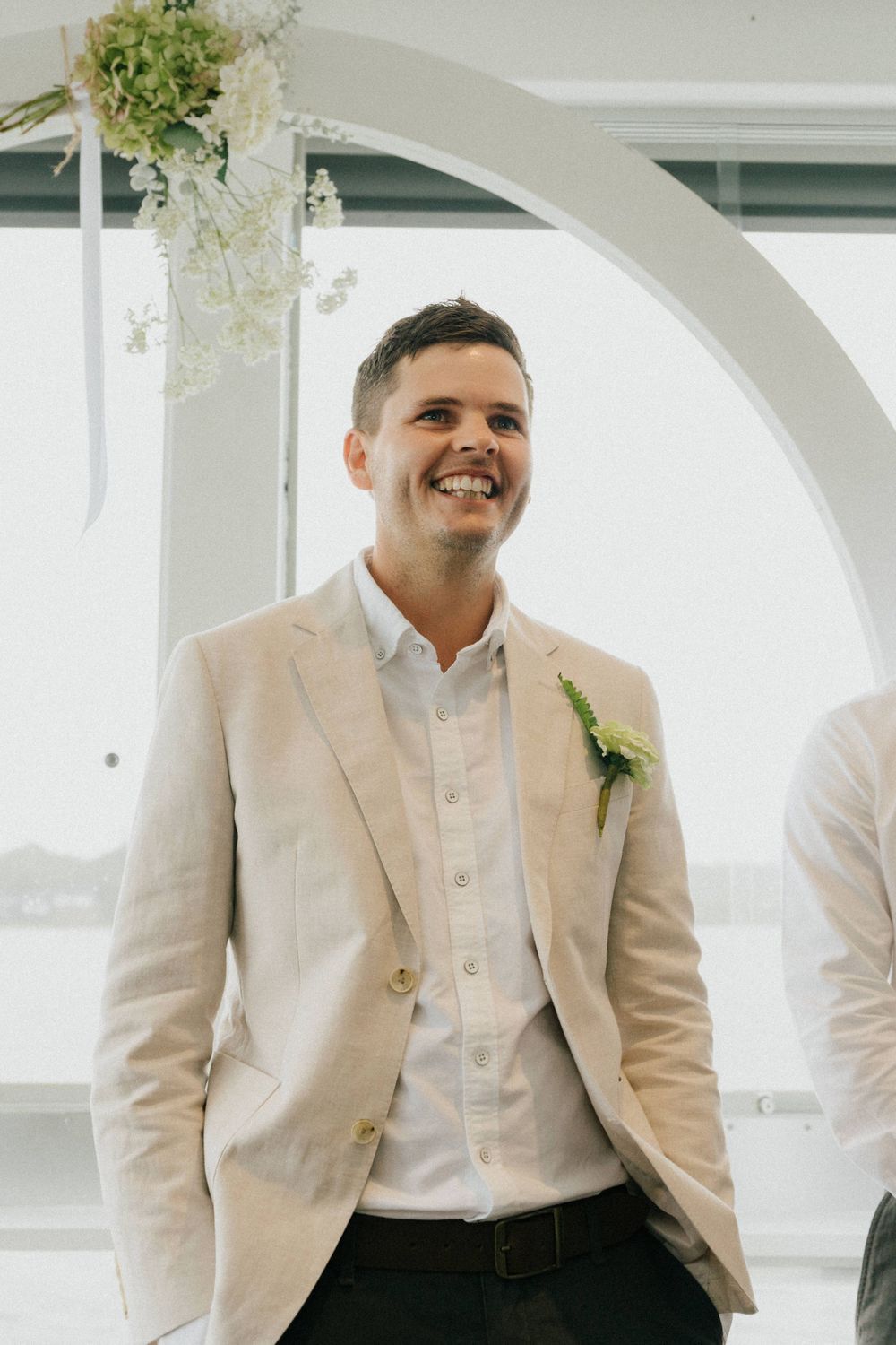Groom in beige suit jacket with boutonniere smiling under floral arch decoration.