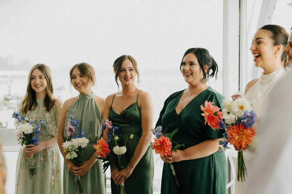 Bridesmaids in emerald green and sage dresses hold colorful bouquets while sharing a candid moment at a wedding.
