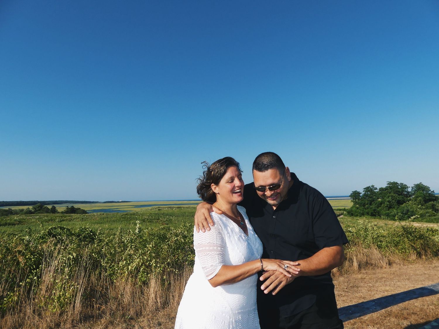 Happy couple smiling in a field under a clear blue sky.