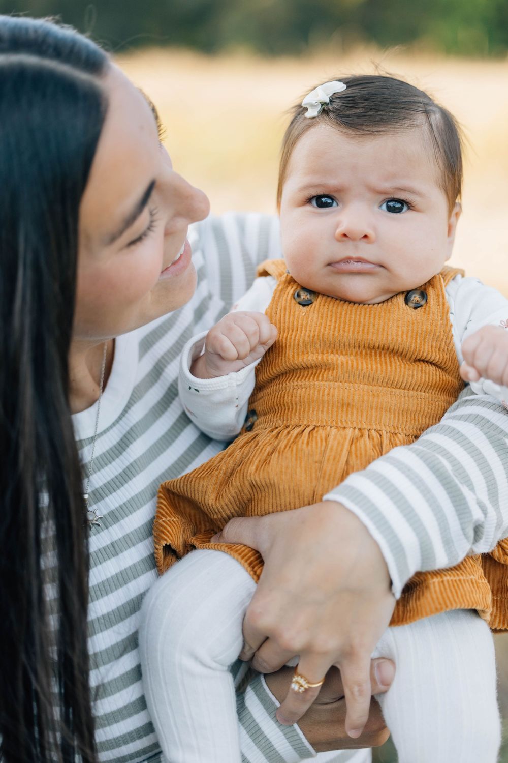 Dark-haired mother gazing lovingly at her baby girl in an orange corduroy dress.
