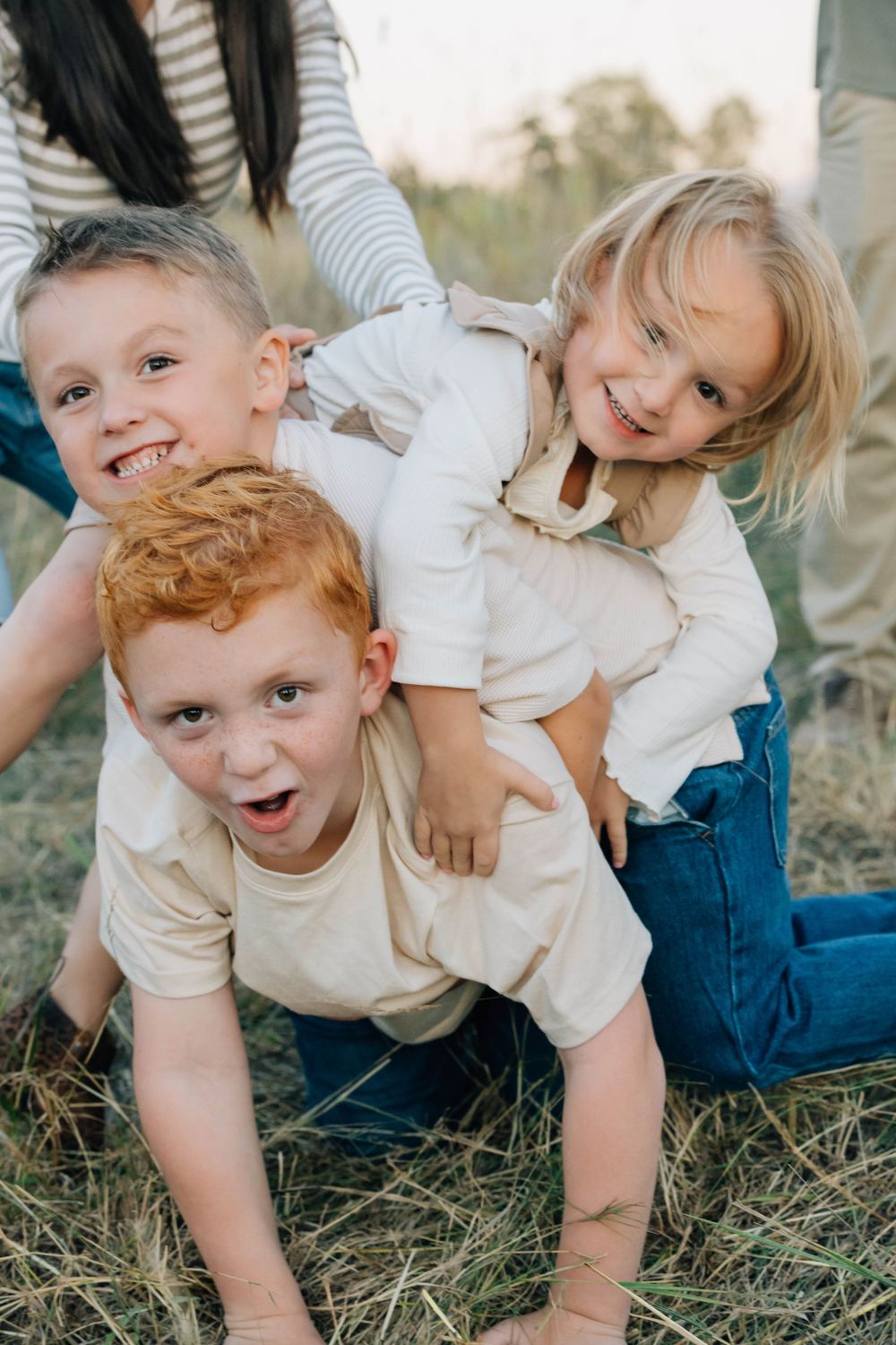 Three young siblings playing and laughing together in a grassy outdoor field.