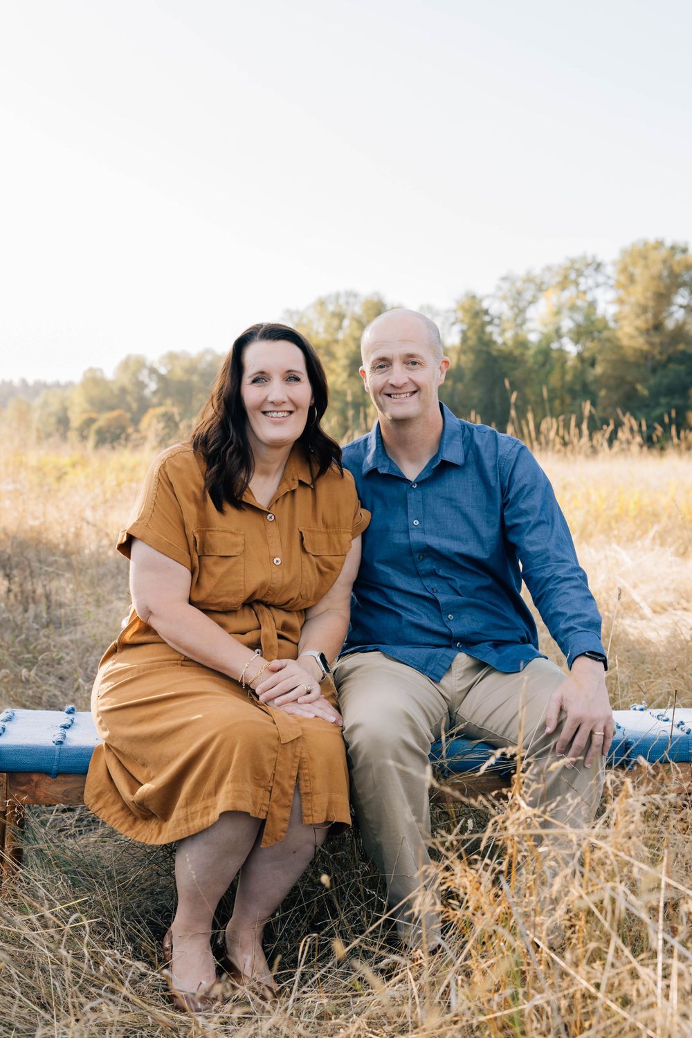 Smiling couple sitting together on a bench in a golden field at sunset.