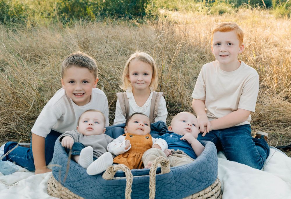 Three older siblings sitting with three babies in a round basket outdoors.