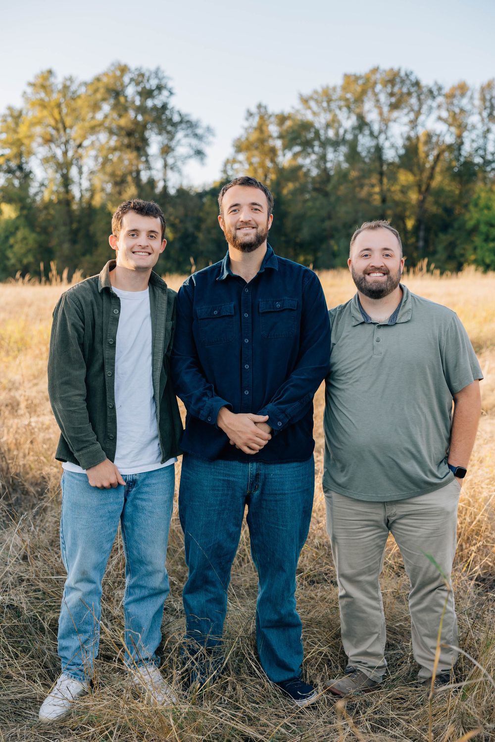 Three young men smiling together while standing in a golden grassy field.