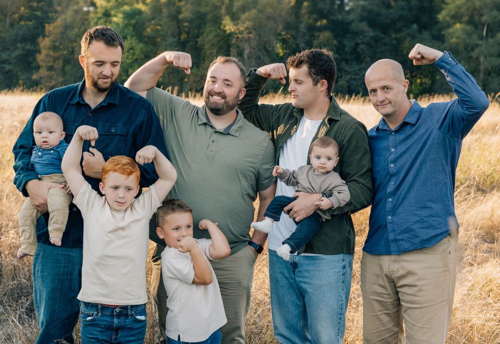 Group of men and children flexing muscles and laughing in a sunny field.