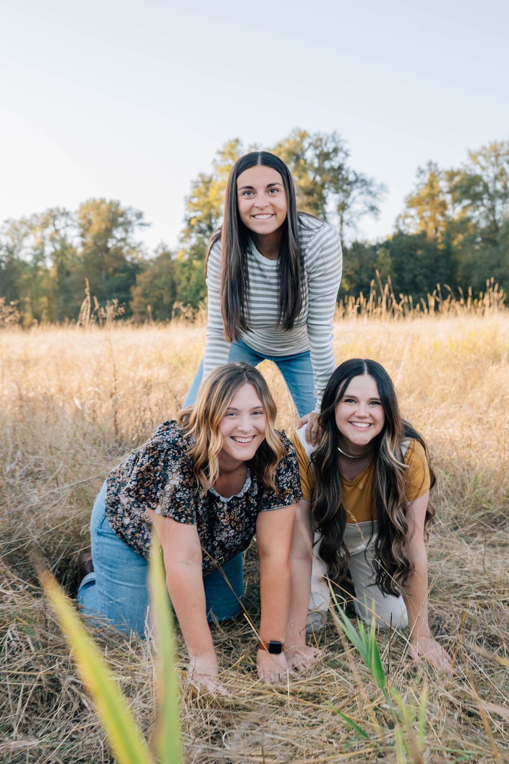 Three smiling sisters posing playfully together in a golden autumn field.
