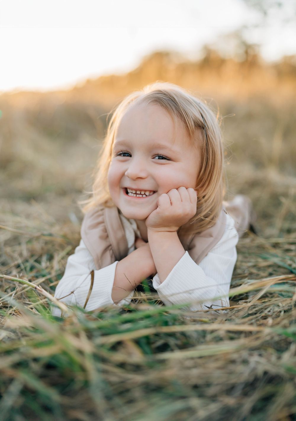 Smiling blonde girl lying in golden grass field, resting chin on hands at sunset.