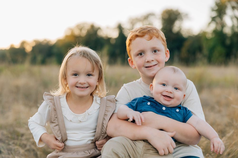 Three young siblings smile together in a golden sunlit field, older boy holding baby with blonde girl beside him.
