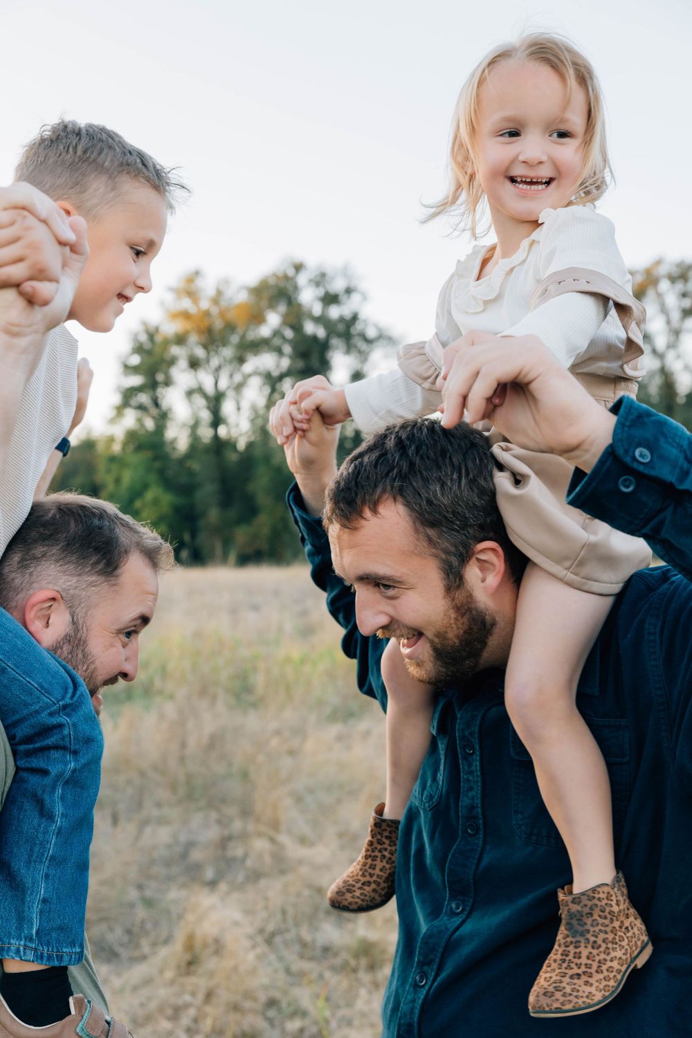 Two dads laughing outdoors as they carry their young children on shoulders and backs in a sunny field.