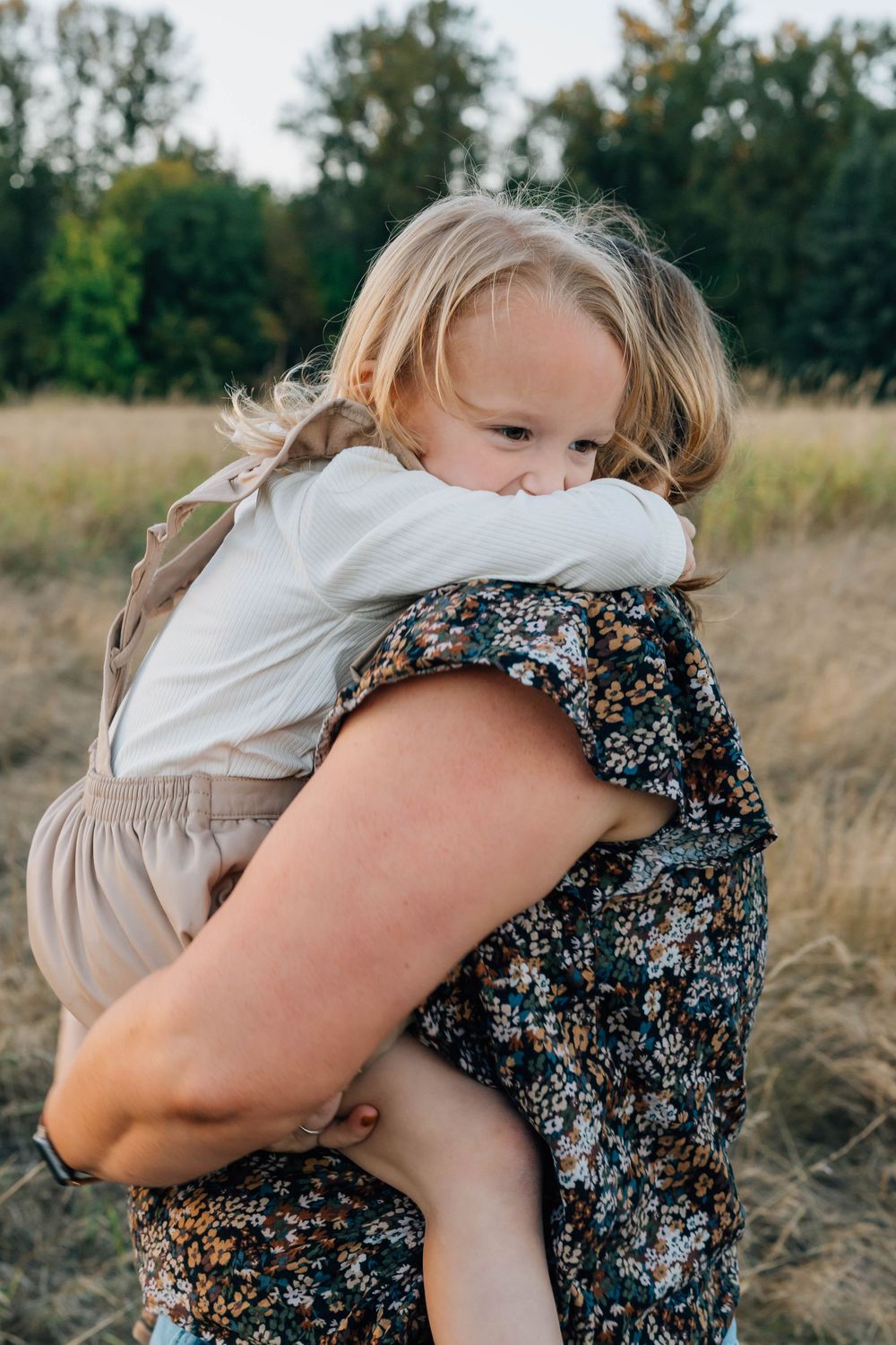 A young blonde toddler hugs an adult wearing a floral dress during an outdoor family photo session.