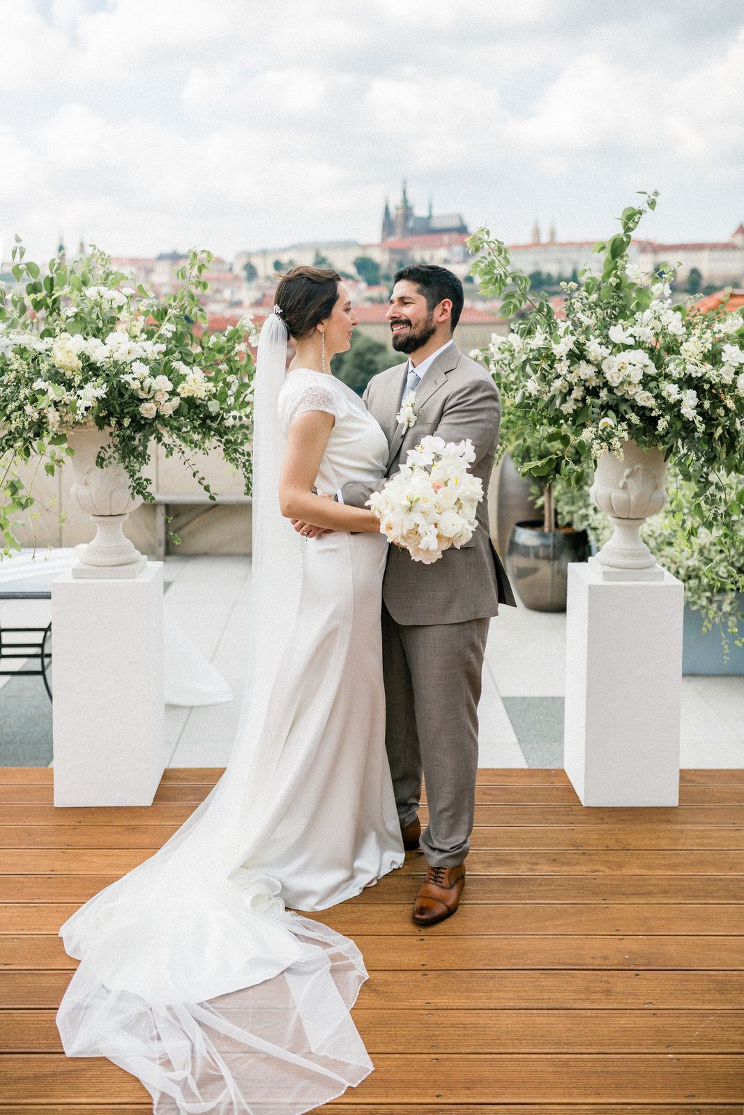 Wedding couple poses on a rooftop terrace with white floral decorations and Prague castle in the background.