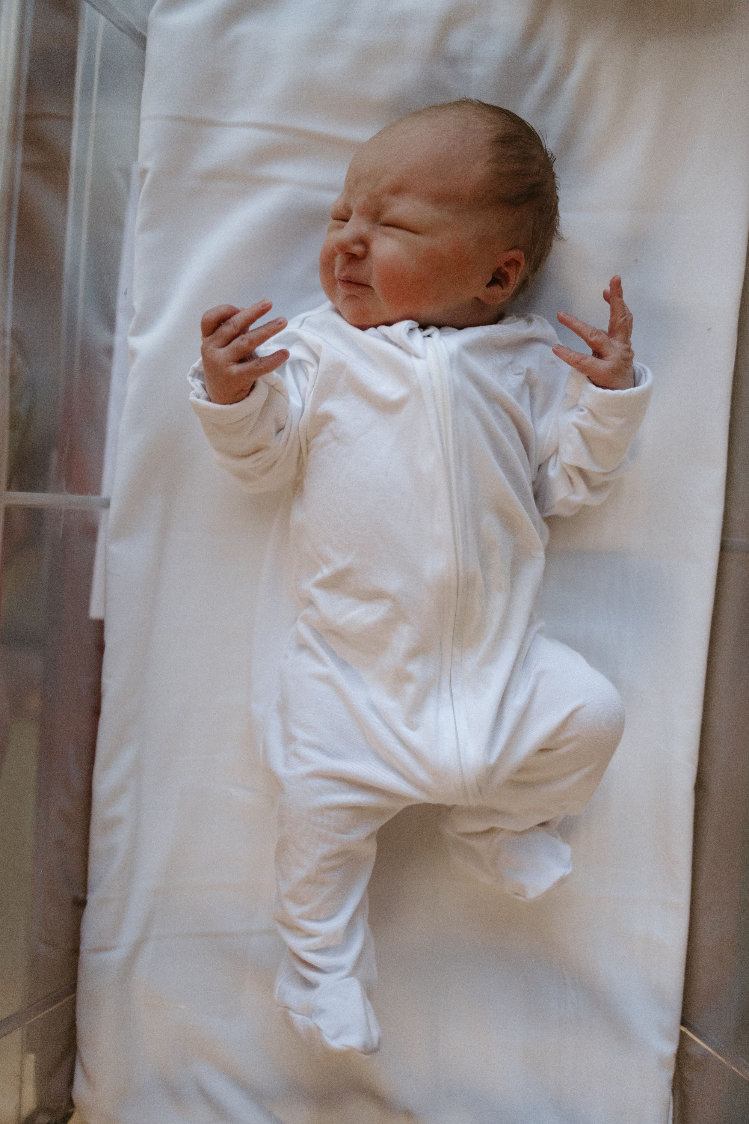Newborn baby in white sleeper stretches on white bedding.