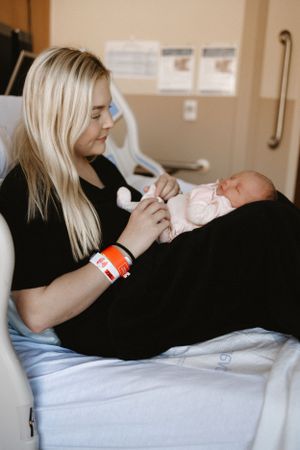 Mother in black shirt holding and feeding newborn baby in hospital room.