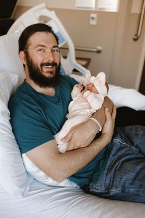 New parent in teal shirt holds sleeping newborn in hospital room.