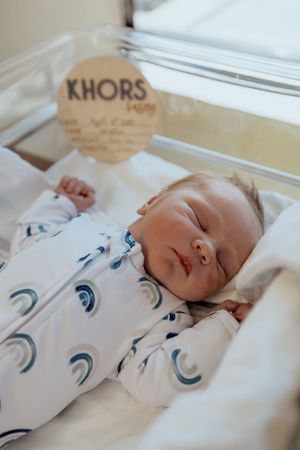 A sleeping newborn baby wearing a white onesie with blue circular patterns lies peacefully on white bedding.