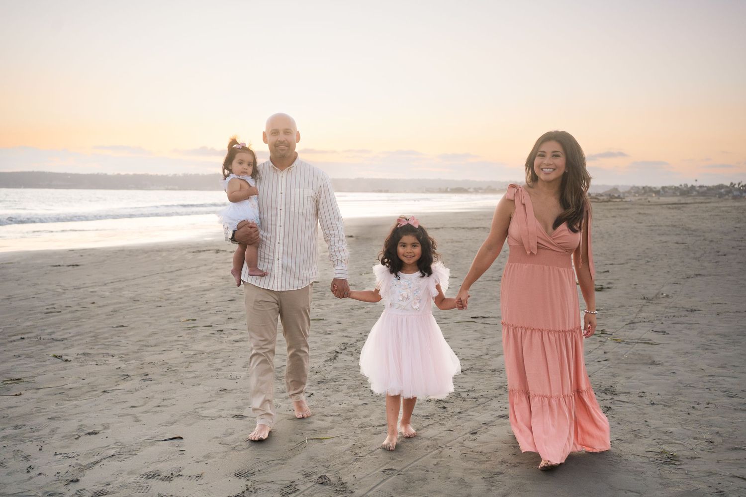 A family strolls together holding hands on a peaceful beach at sunset wearing coordinated pink and white outfits.