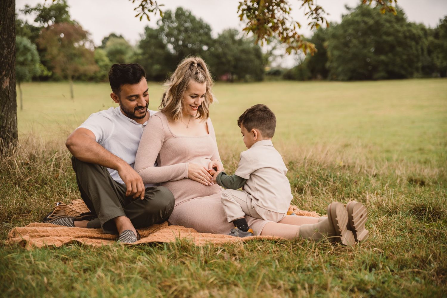 A tender maternity moment shared by a family sitting on a blanket in a grassy field during sunset.