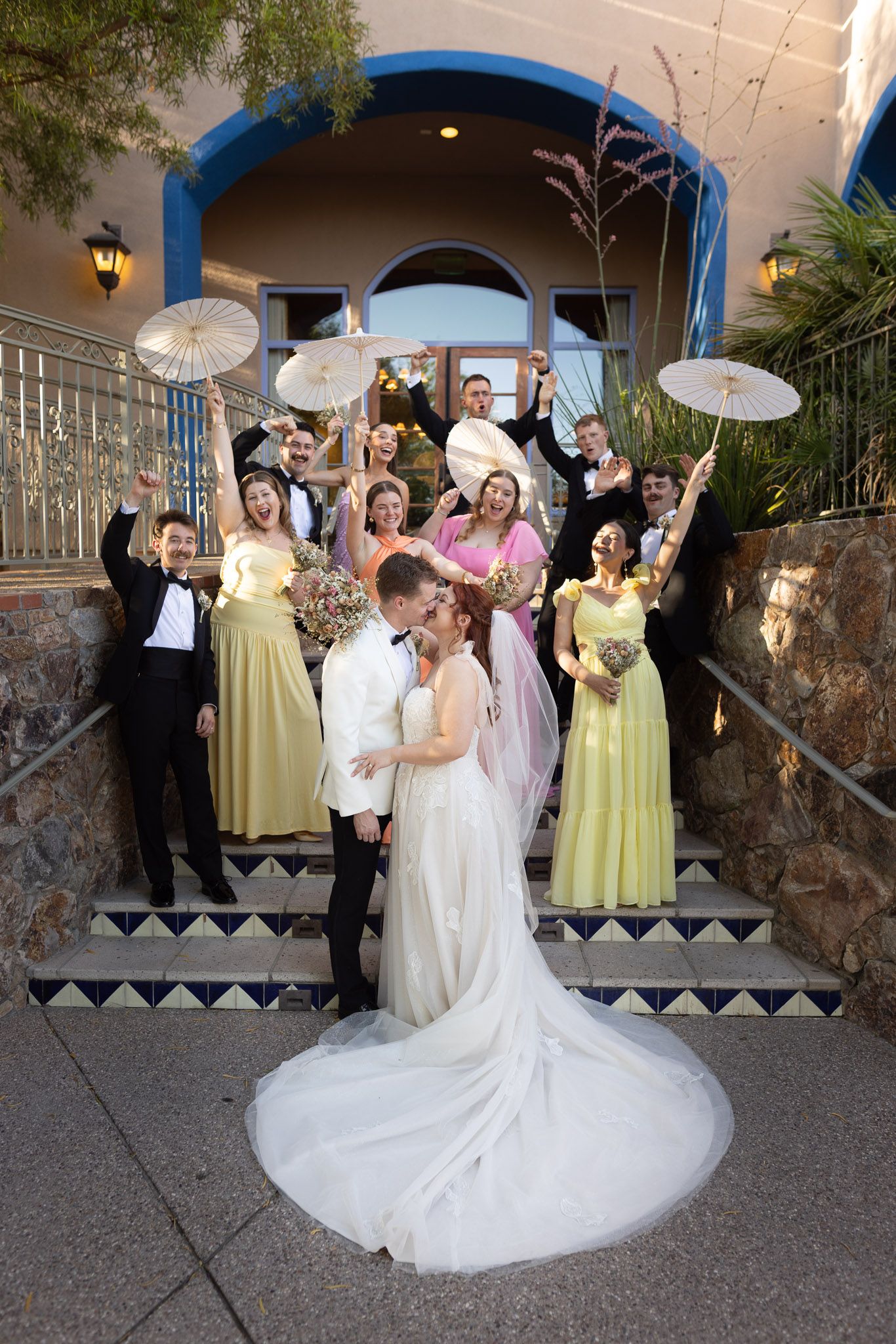 Wedding party celebrating on ornate stone steps with decorative parasols against Spanish-style archway entrance.