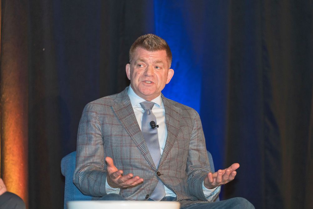 A speaker gestures with both hands while presenting at a podium with dramatic blue lighting in the background.