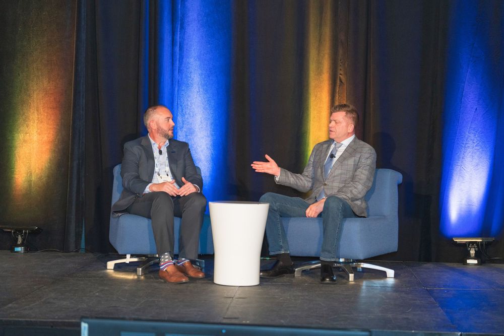 Professional speakers engage in animated discussion on stage at business conference with blue lighting.