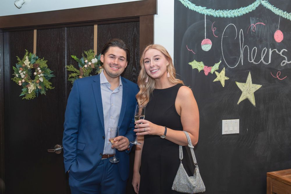 A couple in formal attire poses together at a social event with a chalkboard wall visible in the background.