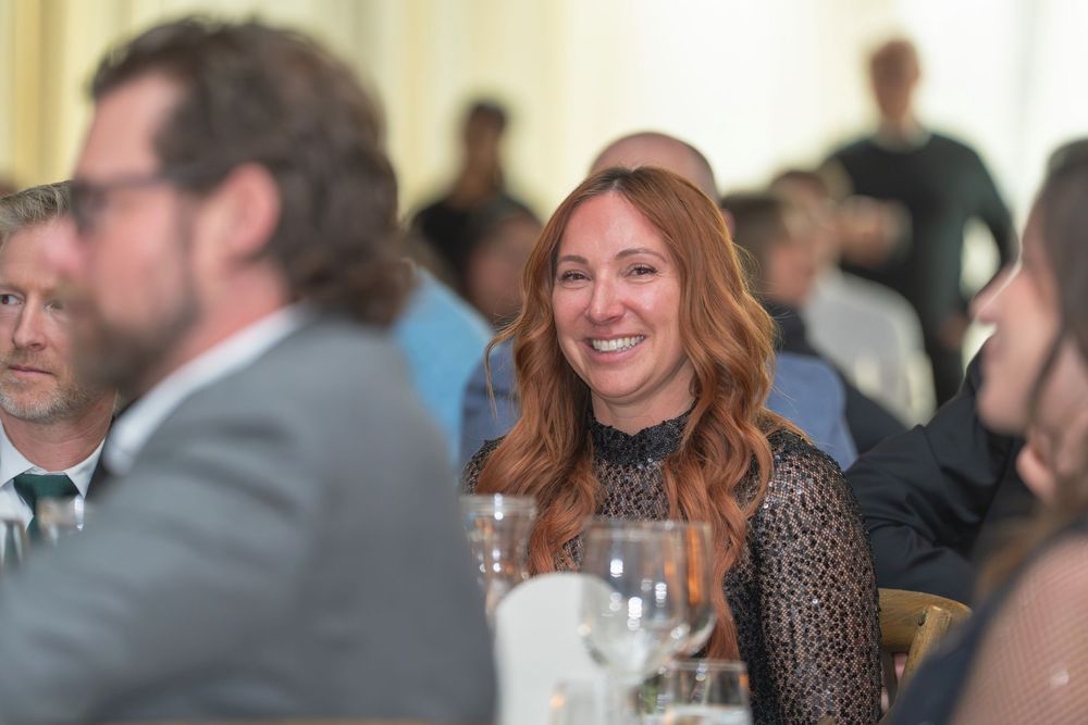 A woman with red hair wearing a dark patterned top smiles while seated at a formal dining event.