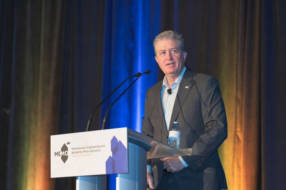 A presenter in a gray suit speaks at a podium with blue and orange stage lighting effects.