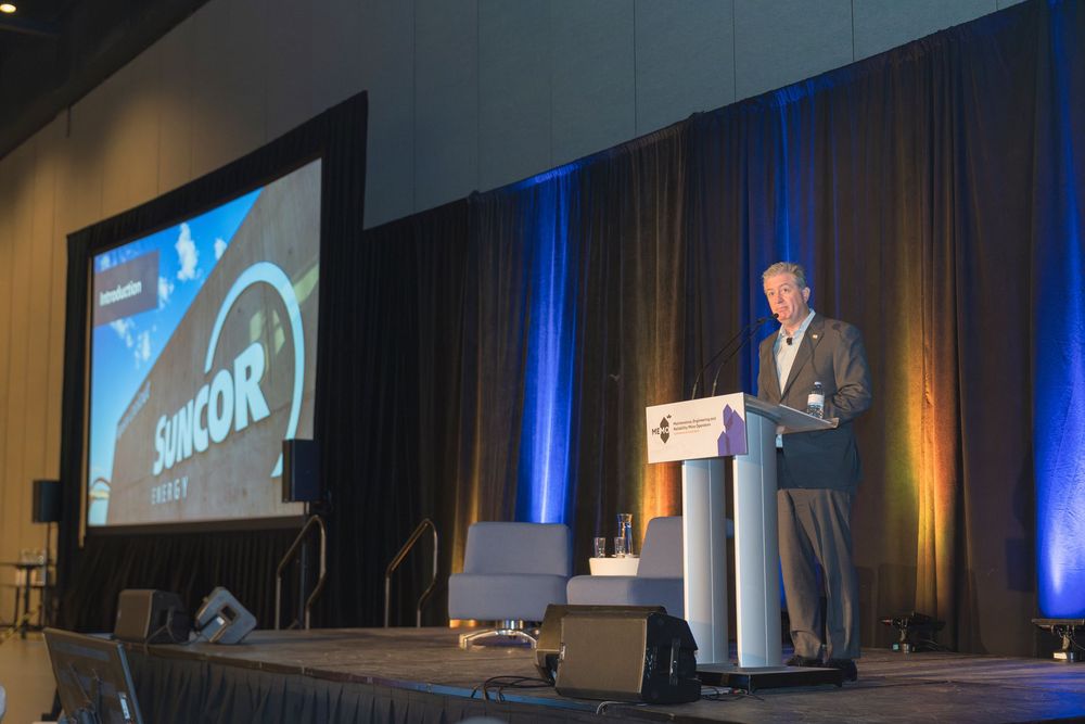 Speaker presenting on stage at a professional conference with large projection screens and blue lighting.
