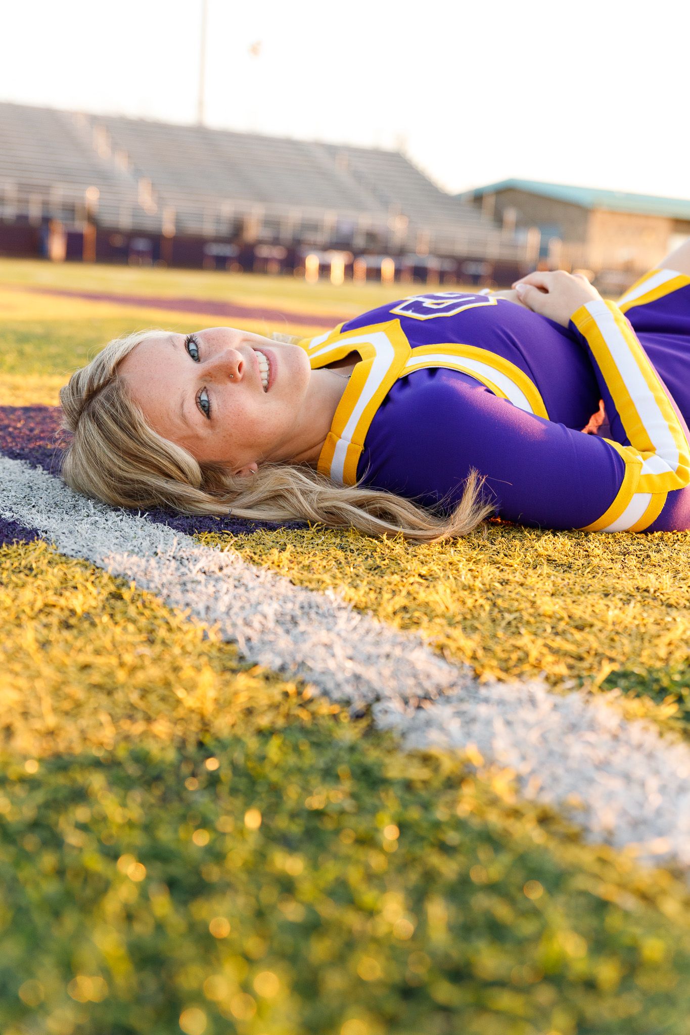 Cheerleader lying on field in purple and gold uniform during golden hour photography session.