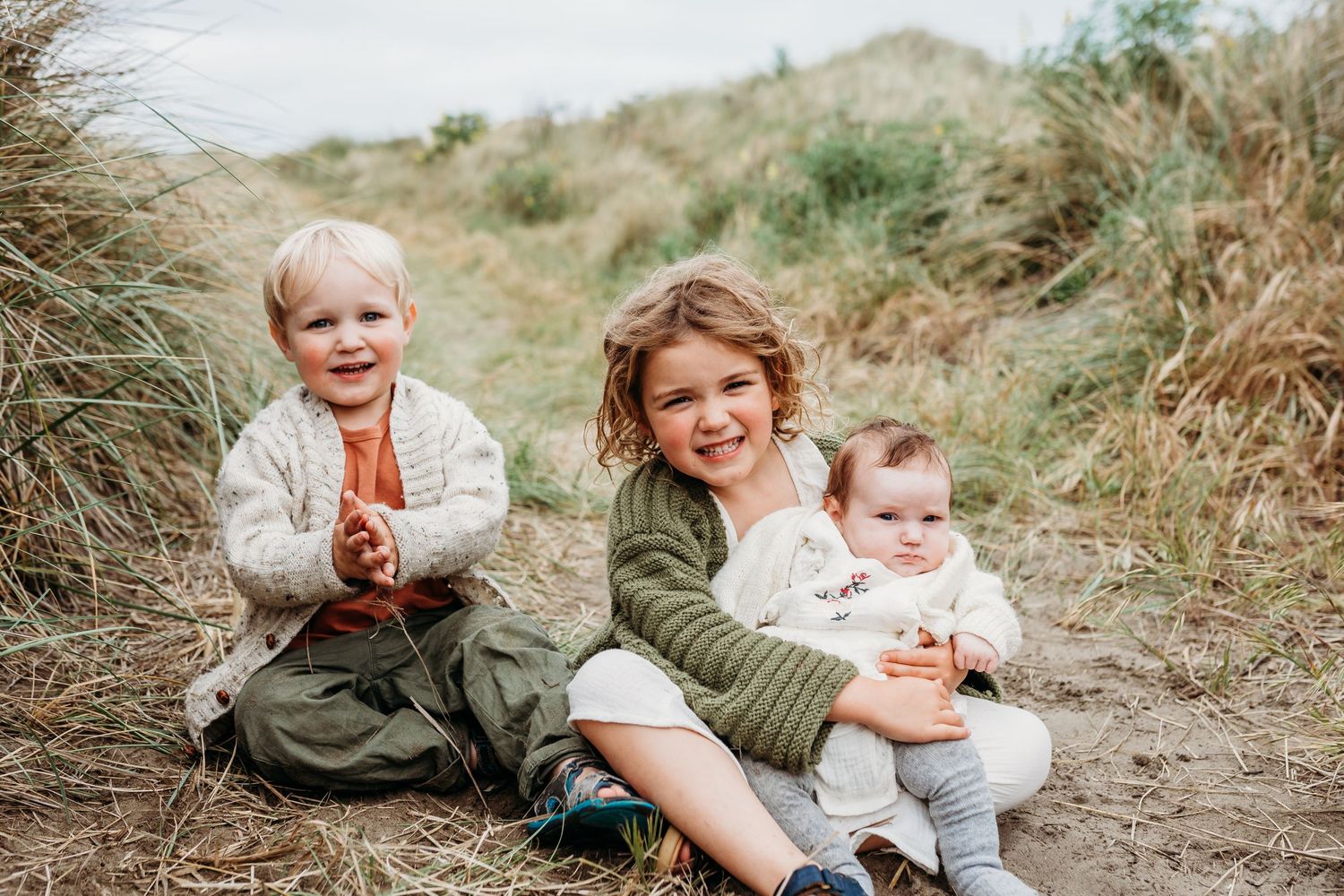 Three young children sitting together on a sandy path among coastal grasses.