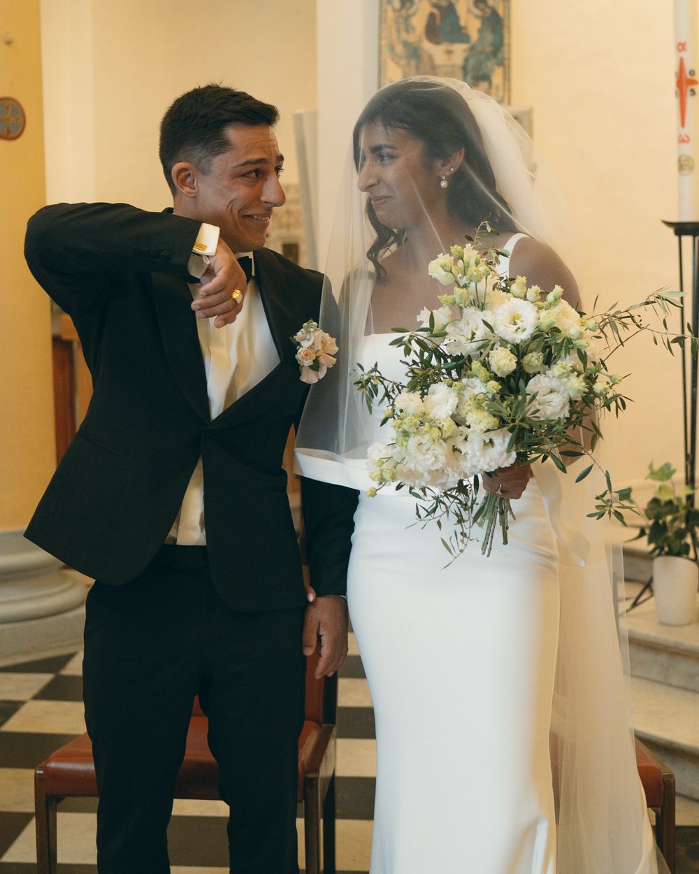Wedding couple in formal attire standing near white floral arrangements in a church.
